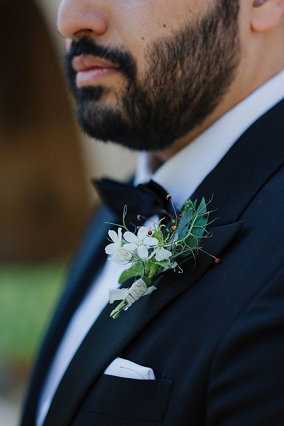 Groom boutonniere with a white flower boutonniere and greenery taped to a black tuxedo lapel, bow tie and pocket square, outdoors blurred greenery