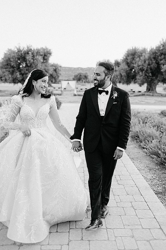 Couple portrait of bride and groom walking hand in hand on a stone walkway, bride in veil and gown looking at him, trees and hills behind