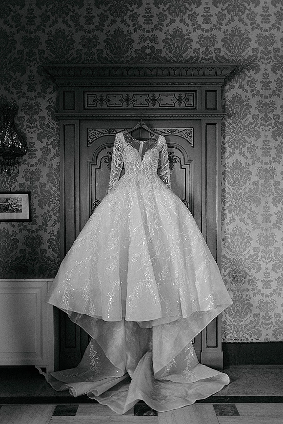Wedding dress, long sleeve wedding dress hanging on a hanger with beaded deep V neckline and train against damask wallpaper and cabinet