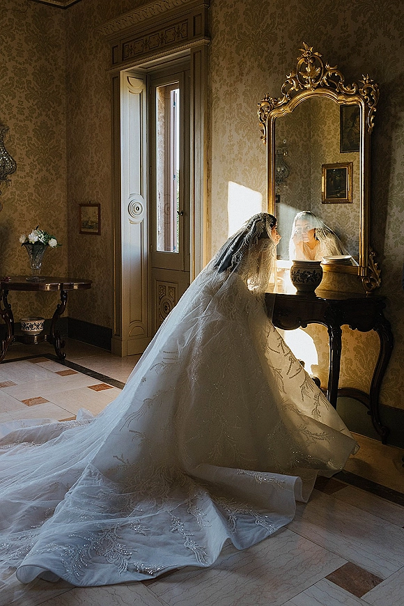 Bridal portrait of a bride in veil with a long train, standing by an ornate gold mirror in a vintage, window-lit room