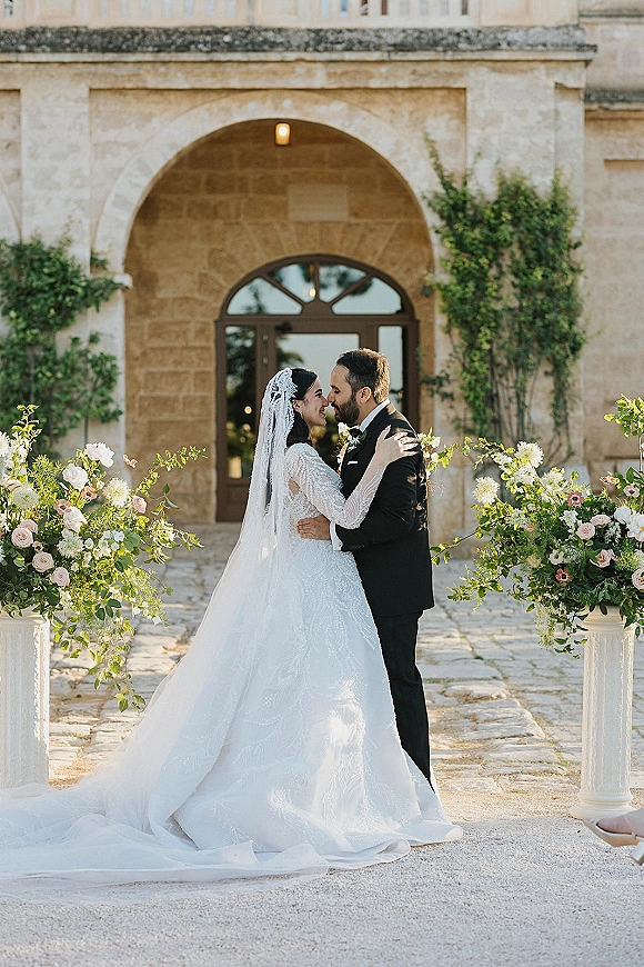 Wedding kiss portrait of bride and groom kissing under a stone archway, her veil draped over lace dress, his tuxedo by pedestal florals
