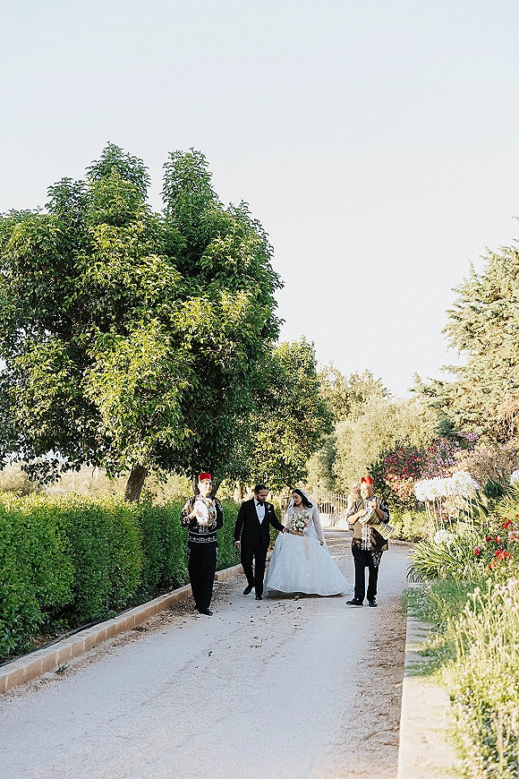 Wedding processional with bride and groom walking hand in hand down a garden path, bride in veil holding bouquet, drummers beside them