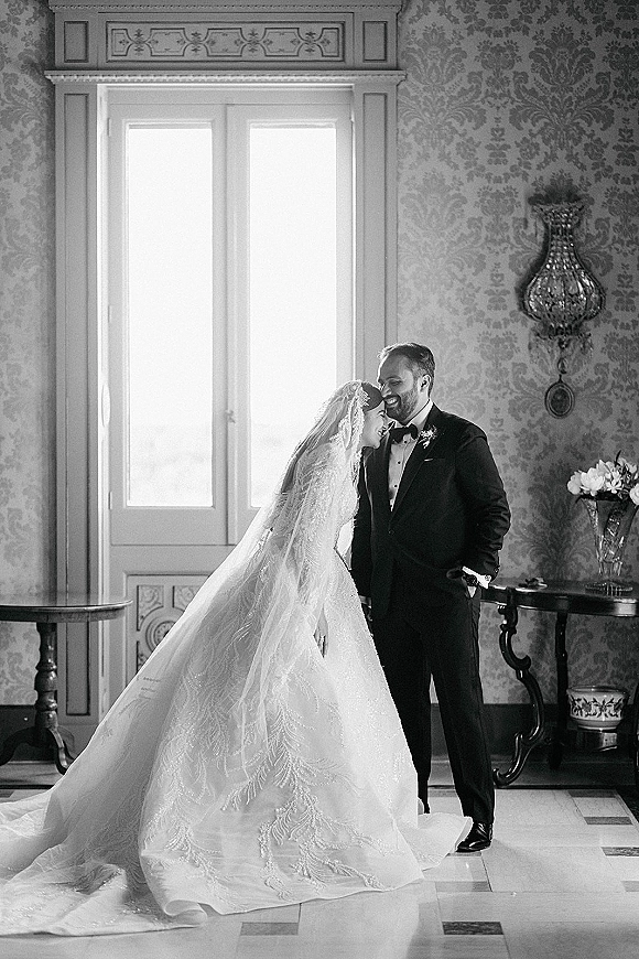 Couple portrait in black and white wedding portrait style, bride laughing and leaning on groom by a large window, veil and lace train flowing indoors