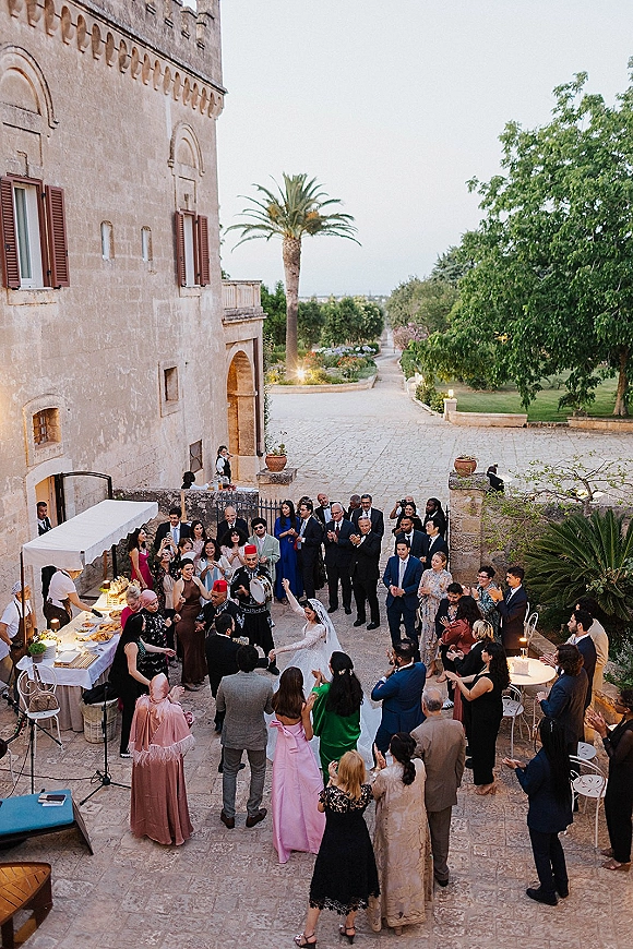 Wedding reception party with guests in a dance circle around the bride in veil and suits, under string lights in a stone courtyard