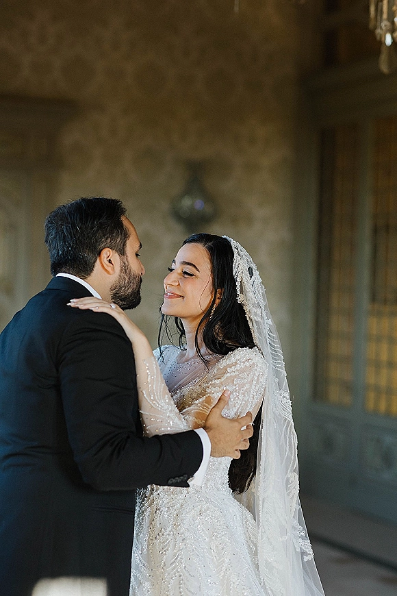 Wedding couple portrait of bride and groom embrace, bride in beaded dress with veil, forehead kiss by stone wall doorway under chandelier