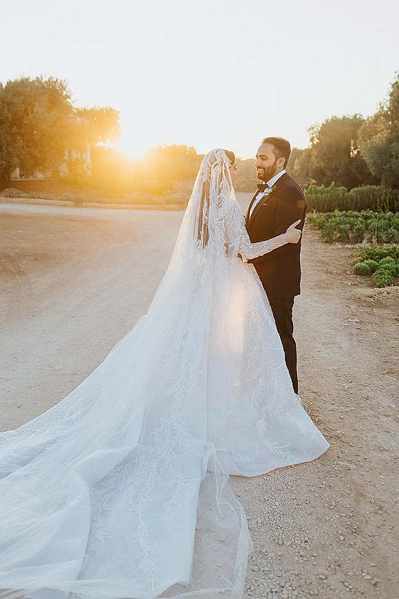 Couple portrait of bride and groom embrace on a dirt road at sunset, her lace long-sleeve gown and cathedral veil backlit by golden sky
