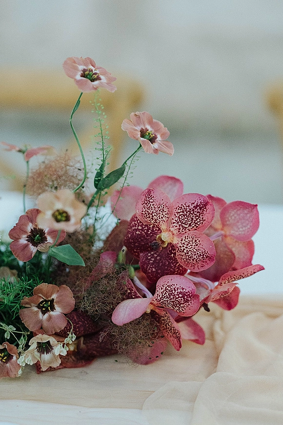Wedding bouquet with pink orchids and blush flowers, trailing greenery and beige ribbon on a neutral tabletop backdrop