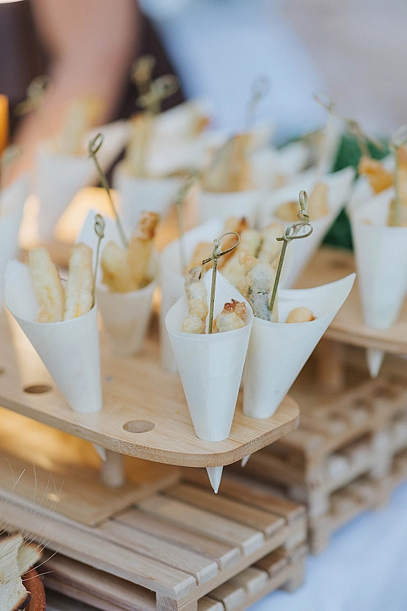 Wedding cocktail appetizers in paper cones with fried bites and wooden pick skewers on a wooden stand, with blurred guests behind