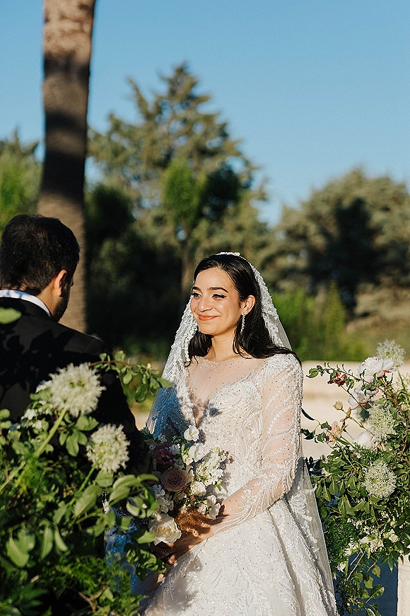 Ceremony moment as bride smiles at groom, holding a white and blush bouquet with long veil and lace dress in a garden setting