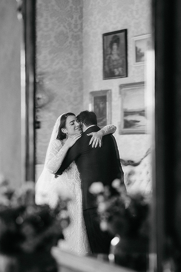First dance as bride and groom dancing in a close embrace, her beaded long-sleeve gown and veil beside framed artwork in a reception room