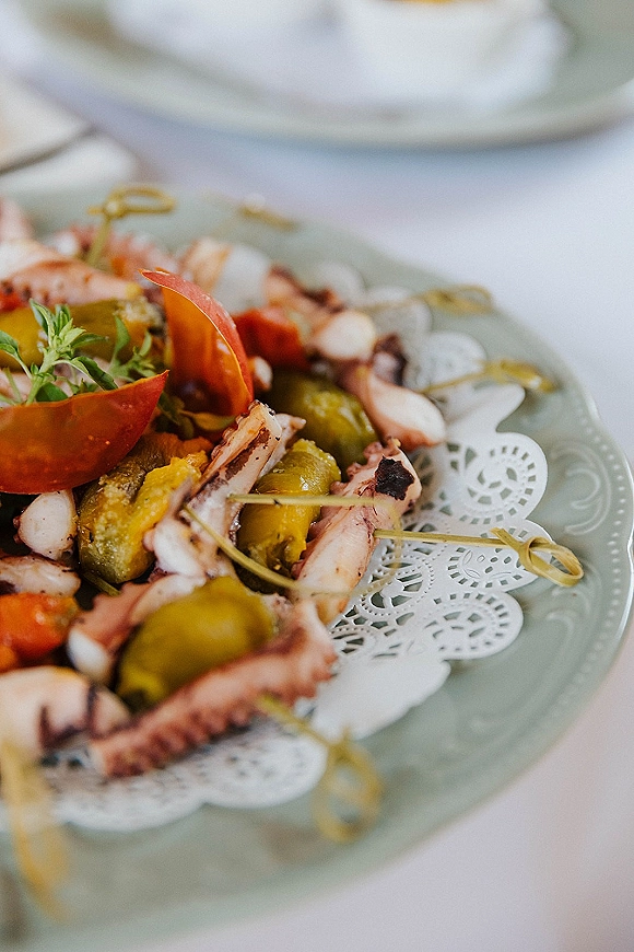 Wedding cocktail appetizers arranged as octopus, olive, and tomato skewer bites on a vintage-style plate with herb accents on a tabletop