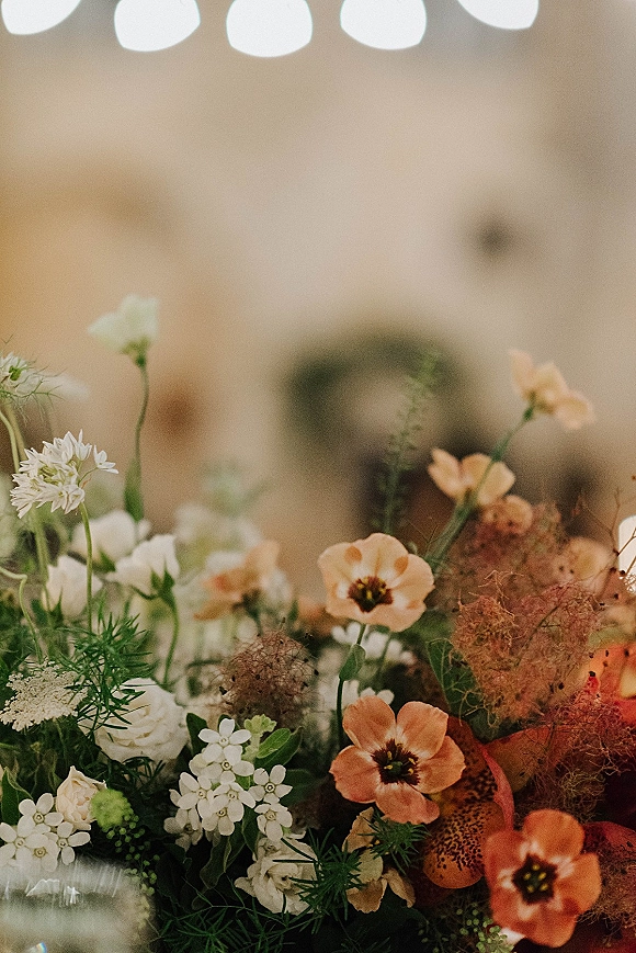 Wedding flowers in a peach and white arrangement with lush greenery, set against a softly blurred indoor venue with arched windows