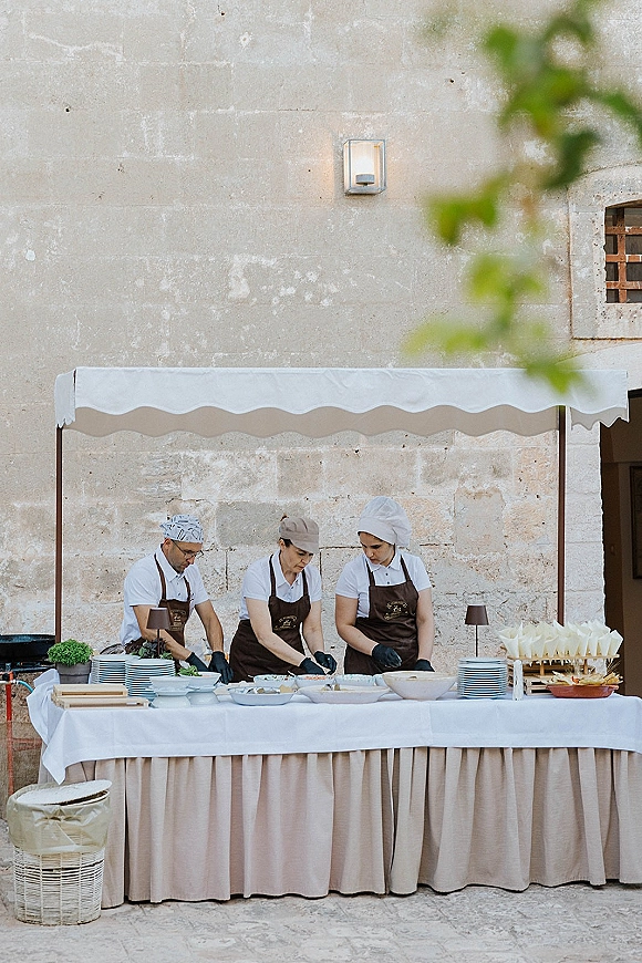 Wedding catering station with an outdoor buffet setup under a canopy, skirted white table and appetizer cones beside a stone wall patio backdrop