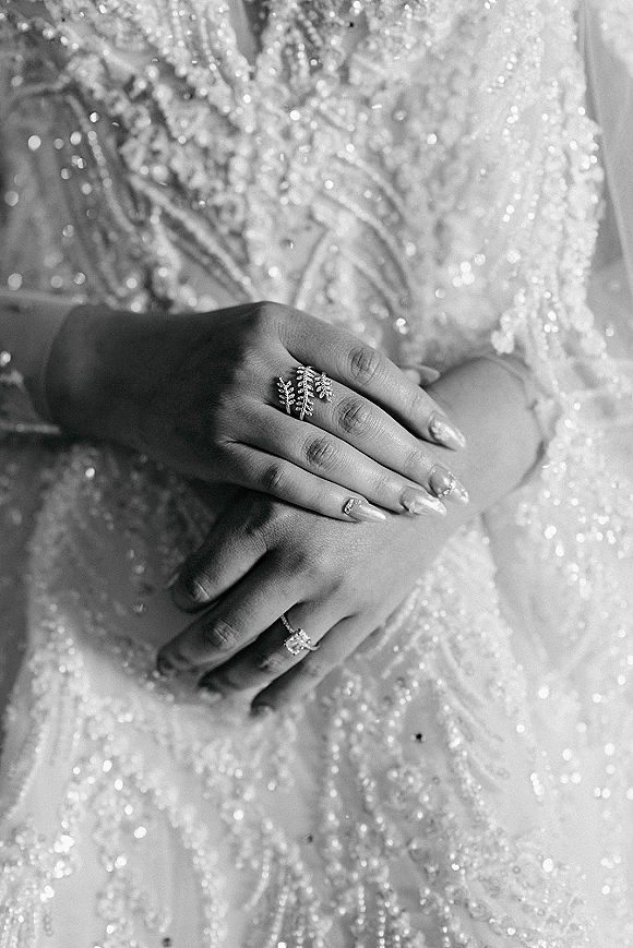 Wedding ring close-up with engagement ring close up on manicured hand, leaf band and bridal set resting against sparkly beaded dress fabric