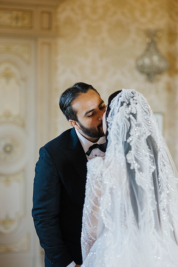 Wedding kiss as bride and groom kiss under a lace veil, her embellished gown and his bow tie framed by patterned wallpaper and sconce