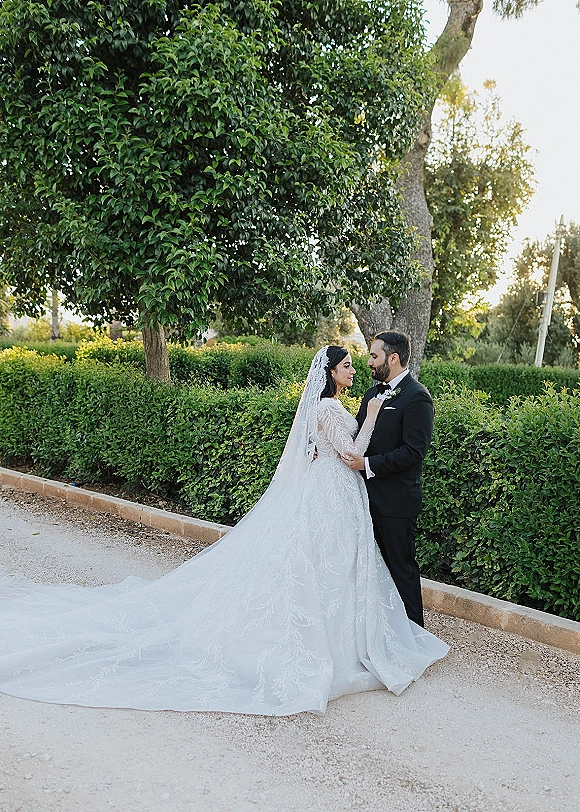 Couple portrait of bride and groom embracing, her long sleeve lace gown and cathedral veil beside his tuxedo on a garden path