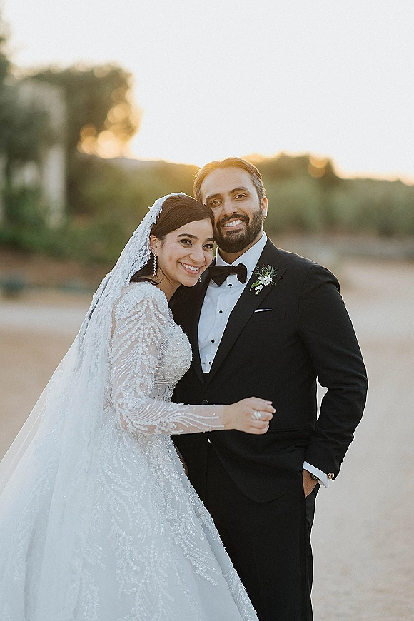 Couple portrait of newlyweds hugging, bride leaning on groom in beaded dress and lace veil, with sunset hills and trees behind