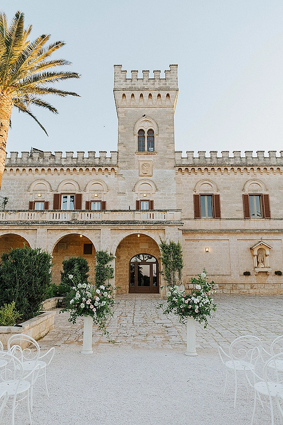 Outdoor ceremony setup in a wedding ceremony courtyard with pedestal floral arrangements lining an aisle before a stone villa under blue sky