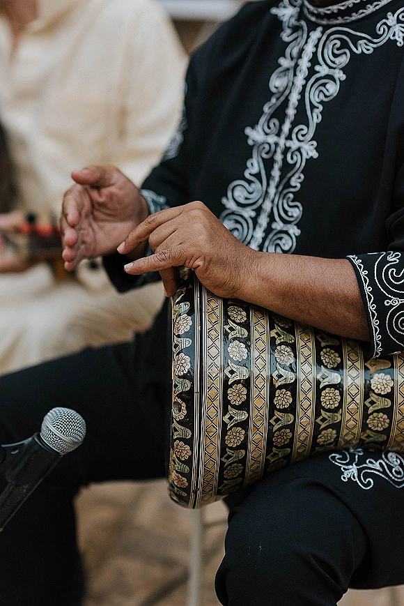 Wedding musician playing hand drum and singing into a microphone, wearing an embroidered outfit with seated guests and chairs behind