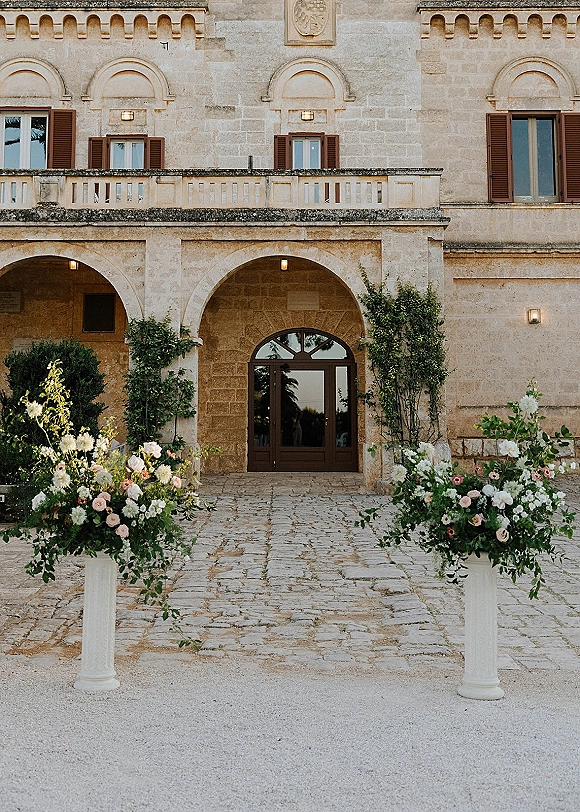 Ceremony entrance decor with wedding entrance flowers on pedestal stands, lush roses and greenery framing an arched stone doorway courtyard