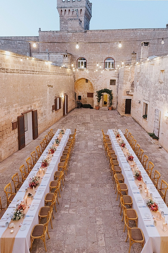 Reception tablescape with long banquet table wedding styling, white linens, floral centerpieces, candles and string lights in a stone courtyard