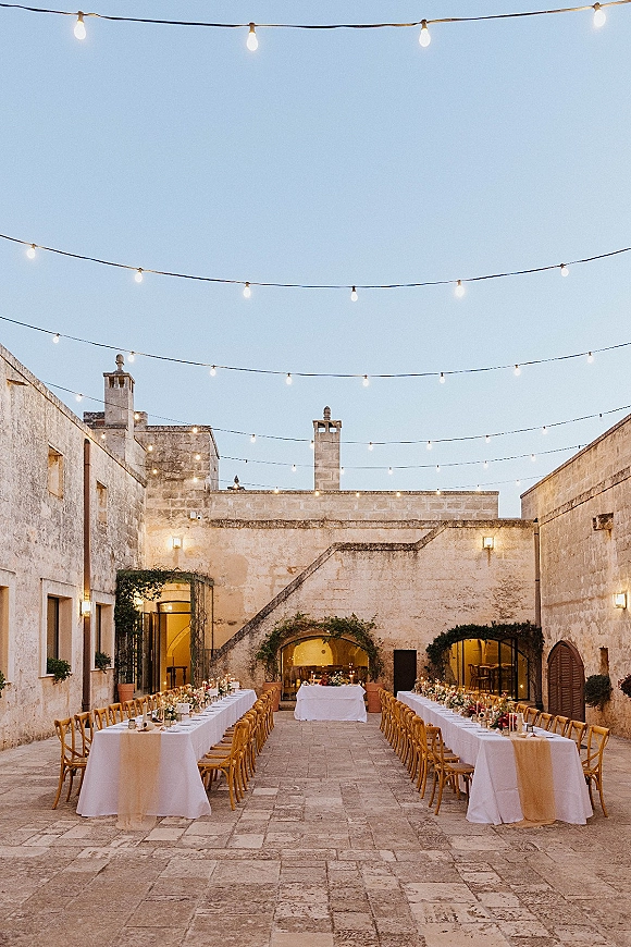 Outdoor reception setup in a courtyard wedding reception with long banquet tables under string lights, candlelit florals in a stone courtyard at dusk