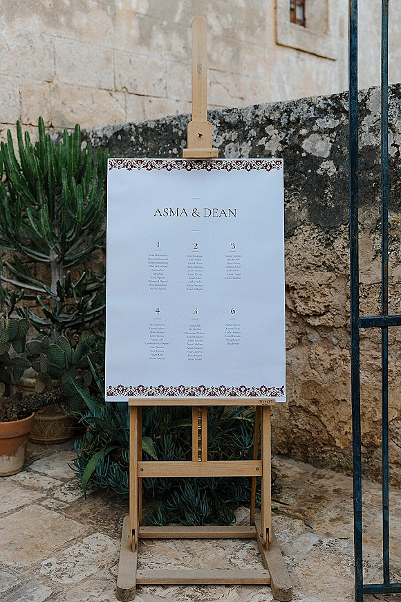 Wedding seating chart on a wooden easel with a patterned border, flanked by potted cactus and succulents in a stone courtyard setting
