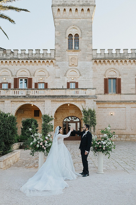 Couple portrait of bride and groom holding hands as she twirls, long veil and ball gown flowing in a stone villa courtyard with floral pedestals