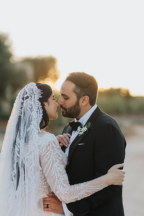 Wedding couple portrait with bride and groom forehead to forehead, lace veil and beaded dress glowing against a golden sunset over water