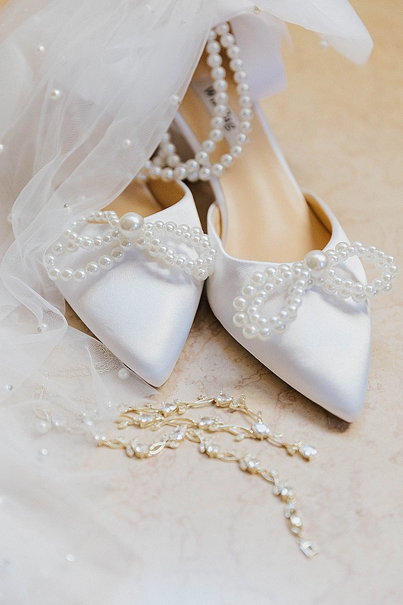 Wedding shoes in white satin with pearl bow shoe clips and ankle straps, arranged with a pearl-accented veil on marble surface