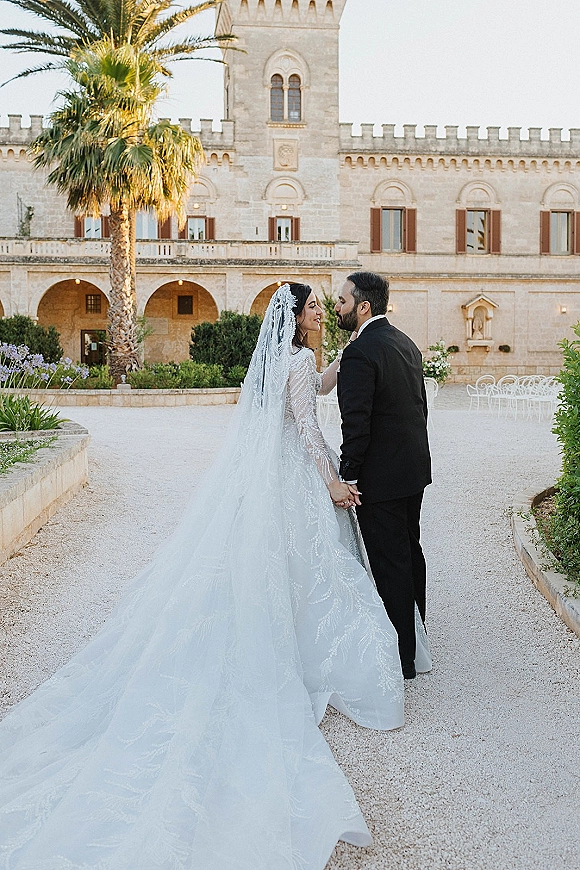 Couple portrait of bride and groom holding hands, gazing at each other in a stone villa courtyard with arched colonnade, lace gown and long veil