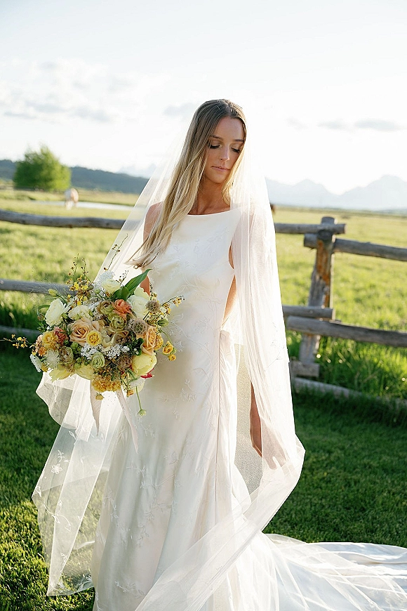 Bridal portrait of a bride holding bouquet with a long veil, in a meadow by a wooden fence with mountains and trees behind