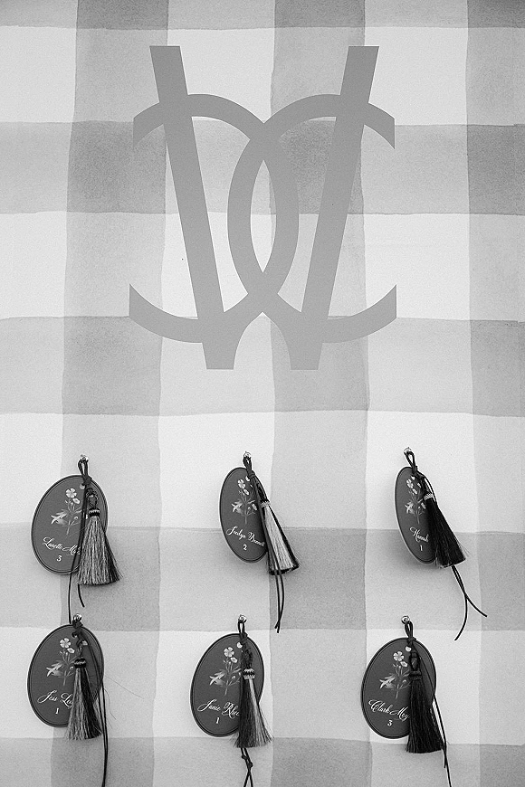 Wedding escort cards with tassels and oval calligraphy tags tied with ribbon, displayed on a gingham fabric backdrop beside a monogram sign