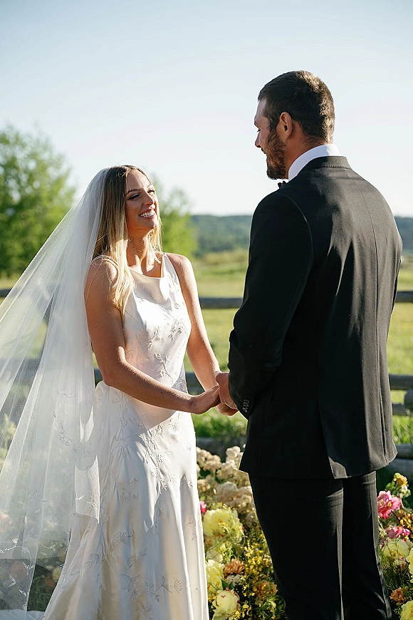 Wedding vows as bride and groom hold hands, her veil draped over embroidered gown beside a wooden fence in a sunny field