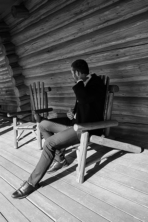Groom portrait of a groom in suit sitting in a wooden rocking chair on a log cabin porch, tie and leather dress shoes visible