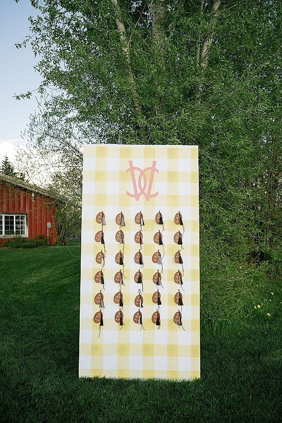 Wedding seating chart with hanging escort card display on a yellow gingham monogram board, set outdoors near a red barn and trees