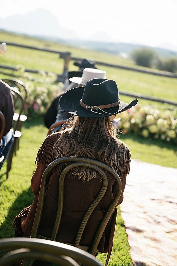 Wedding guest seating with a cowboy hat and fringed jacket on wooden chairs beside an aisle runner, mountain field behind in sunlight