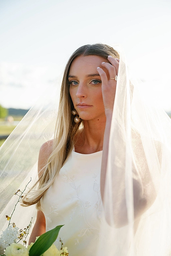 Bridal portrait with a bridal veil draped over her face, holding a greenery bouquet, pearl drop earrings and ring in a field under open sky