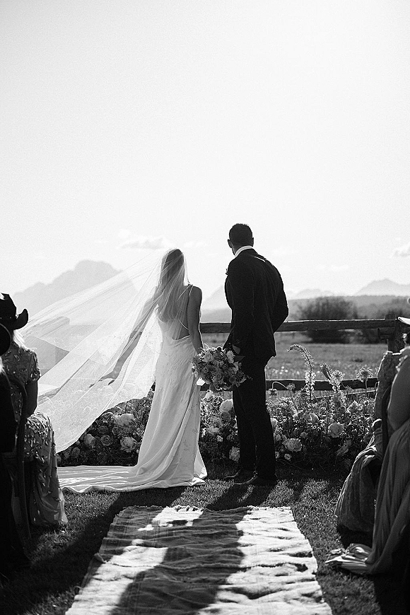 Ceremony moment as bride and groom face the altar, long veil and bouquet beside floral aisle runner with mountains behind guests