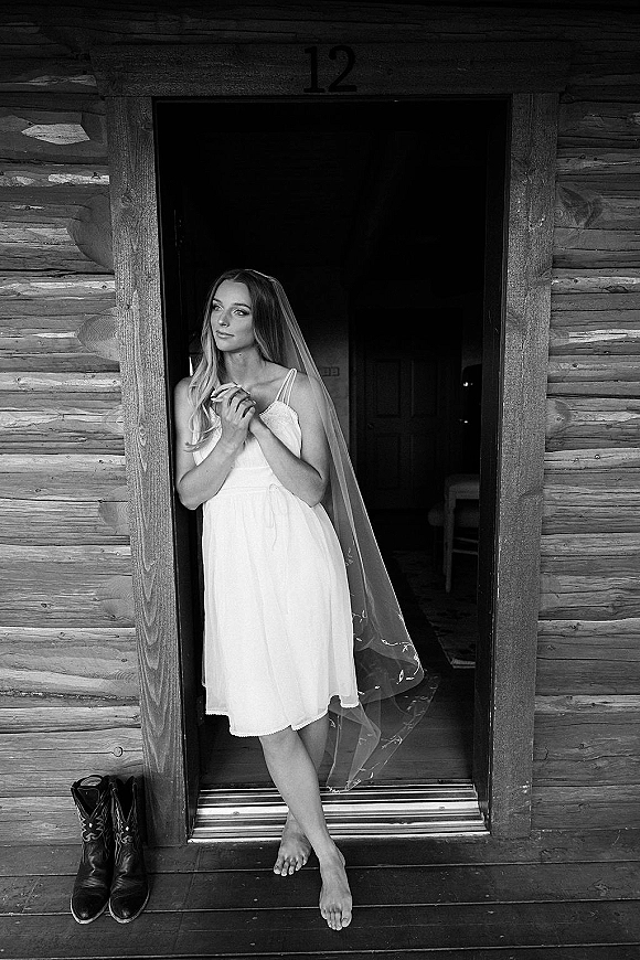 Bridal portrait of a bride in a doorway wearing a short wedding dress and veil, hands clasped, cowboy boots by a log cabin porch