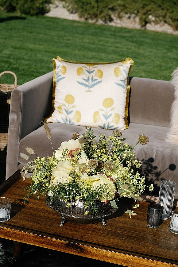 Reception lounge decor with wedding lounge seating featuring a velvet sofa, floral centerpiece, candles, and glasses on a wooden table outdoors