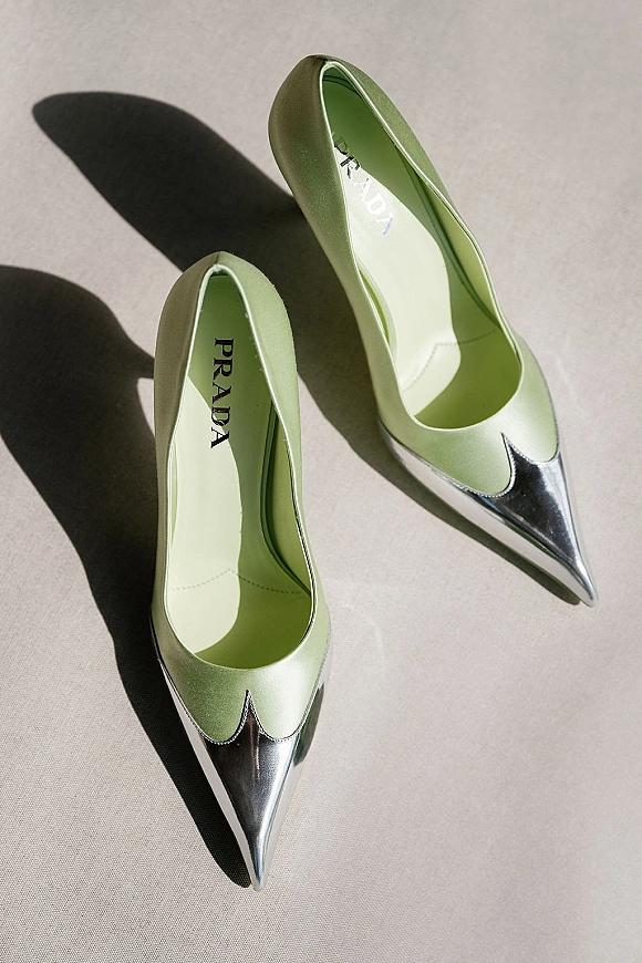 Bridal shoes in sage green heels with metallic silver toe caps, flat lay on neutral fabric with crisp hard shadows