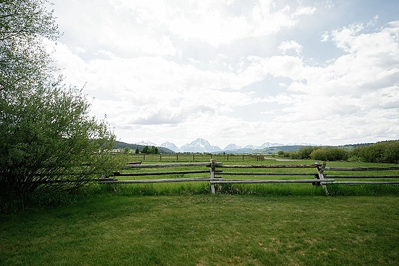 Outdoor landscape with a rustic split-rail fence bordering green meadow grass, shrubs, and trees, with distant mountains under cloudy sky