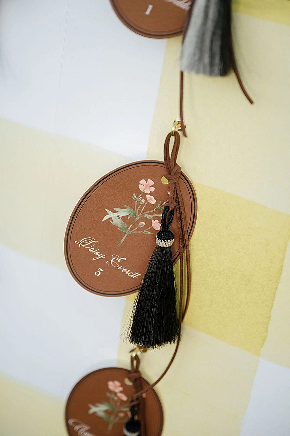 Wedding place cards with round place cards featuring floral illustrations, brass grommets, leather cord, and black tassels on striped fabric