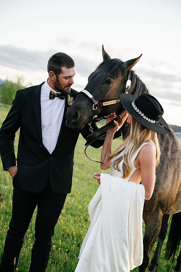 Couple portrait of bride and groom with horse, bride in wedding dress and cowboy hat beside groom in black suit in a grassy field under cloudy sky