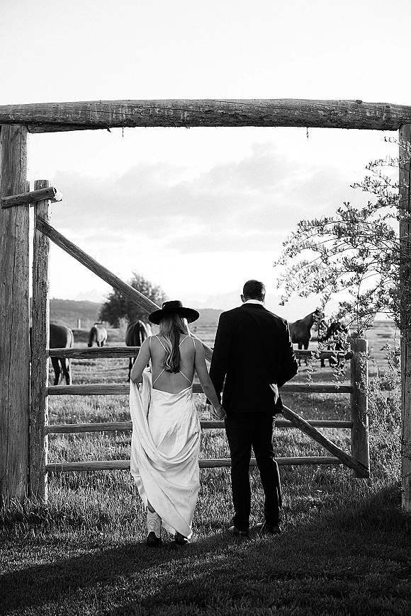 Couple portrait of bride and groom from behind holding hands, bride in cowboy boots and wide brim hat by a wooden gate with horses in pasture