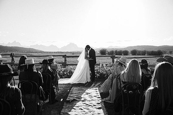 Ceremony kiss at an outdoor wedding ceremony, bride in long veil and groom in black suit on aisle runner with mountains beyond