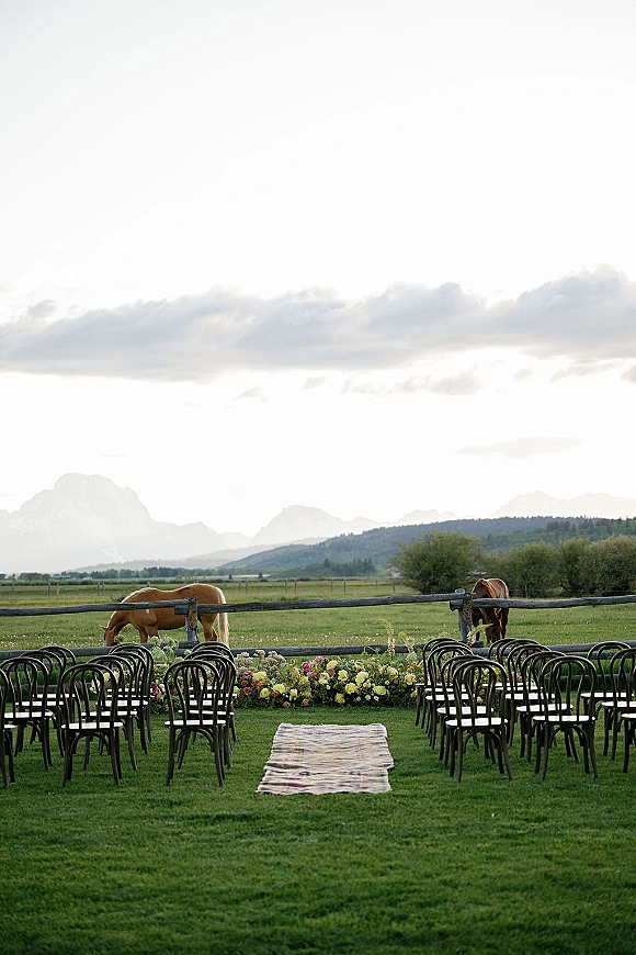 Outdoor ceremony setup with a ceremony aisle runner, chairs and colorful floral arrangements on a ranch lawn with horses and mountains beyond
