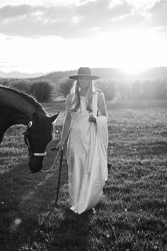 Bridal portrait of a bride in a satin wedding dress and wide brim hat leading a horse across a grassy field at sunset