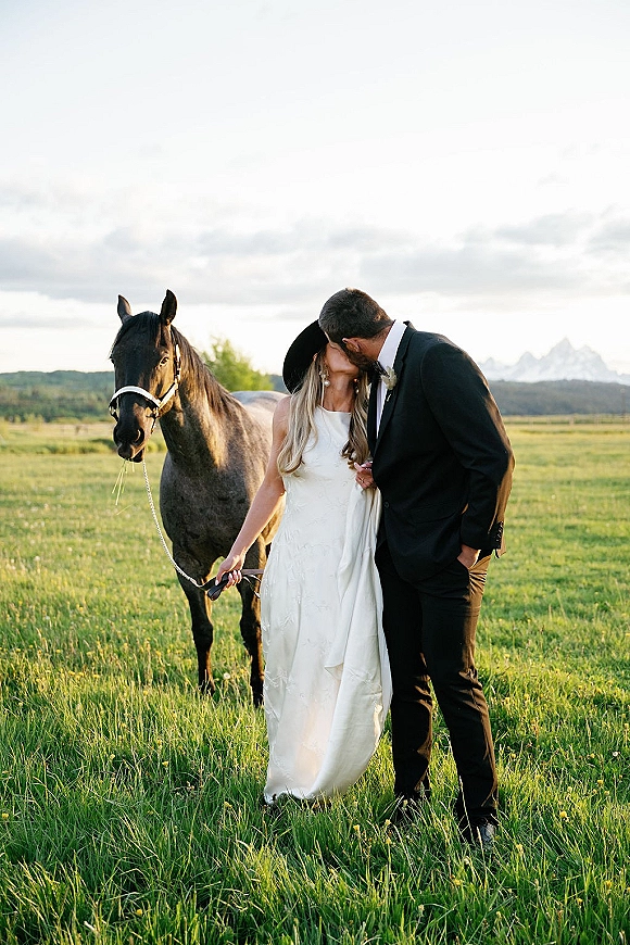 Wedding kiss portrait of bride and groom kissing beside a horse, bride in wide brim hat holding the lead in a mountain field under clouds